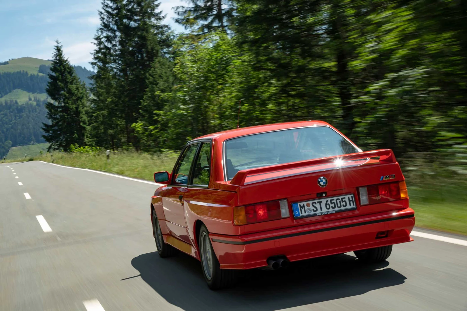 Rdeči BMW M3 E30 med vožnjo, pogled od zadaj na gorski cesti