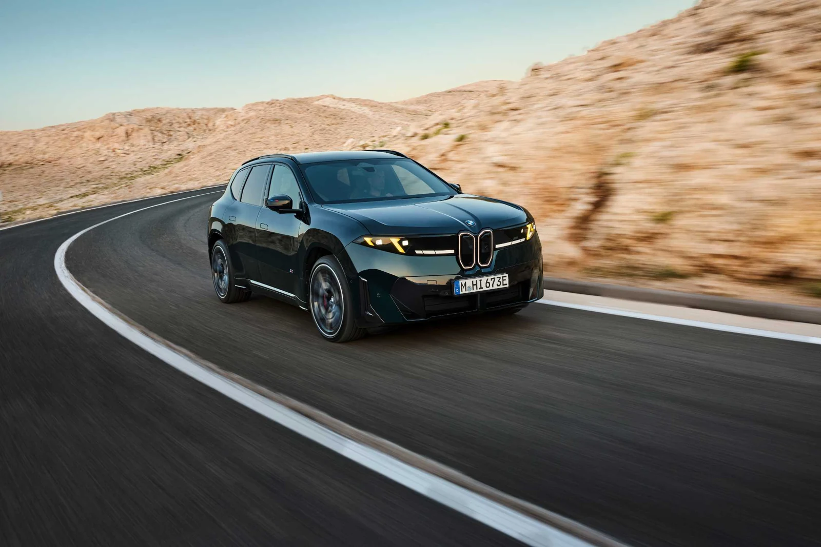 Temnozelen BMW iX3 M60 xDrive z rumenimi svetlobnimi elementi na zaviti cesti ob svetlih skalah