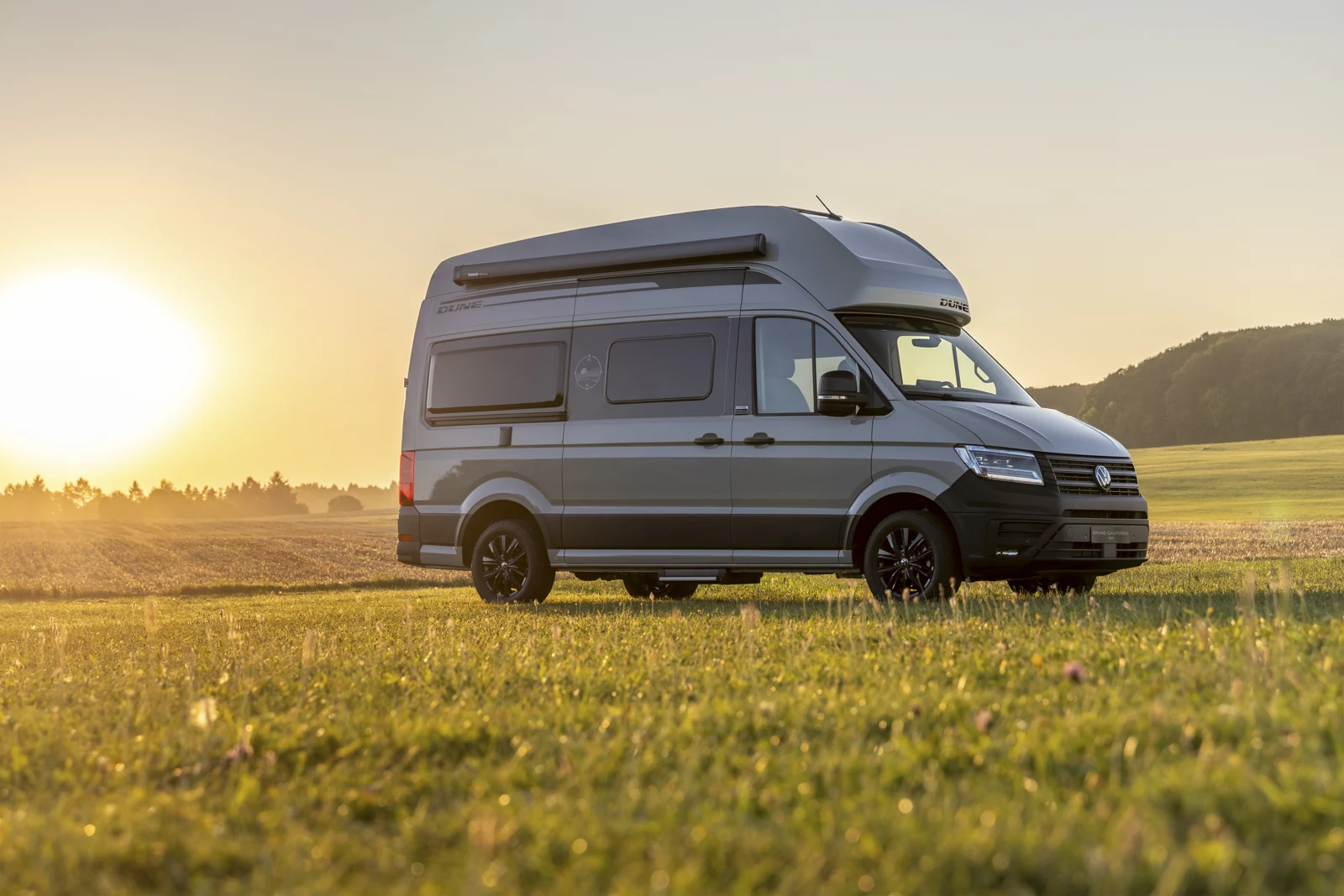 Dejanski siv Volkswagen Grand California Dune, pogled od spredaj s strani na travnati površini ob sončnem zahodu.